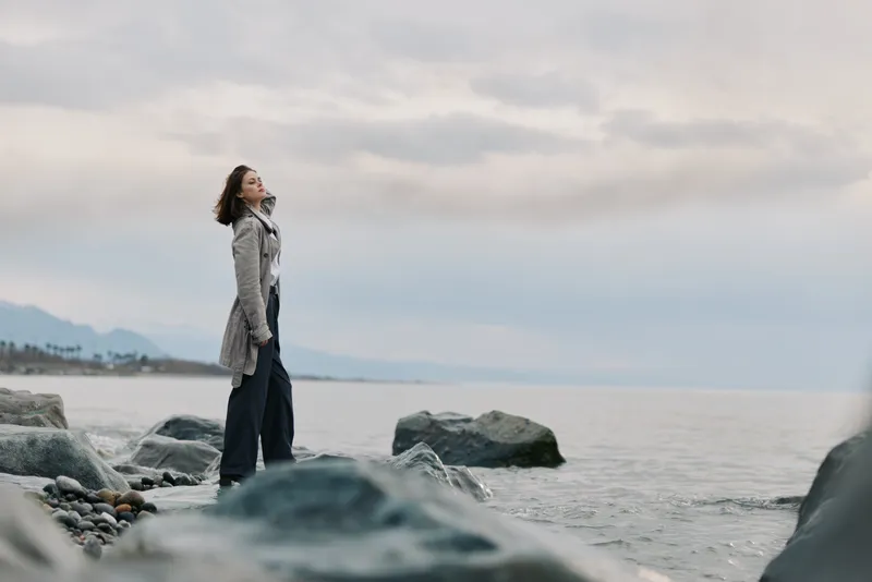 A woman in a long coat stands on rocky shore gazing at calm sea under cloudy sky. The moody atmosphere highlights solitude and contemplation by the water landscape.