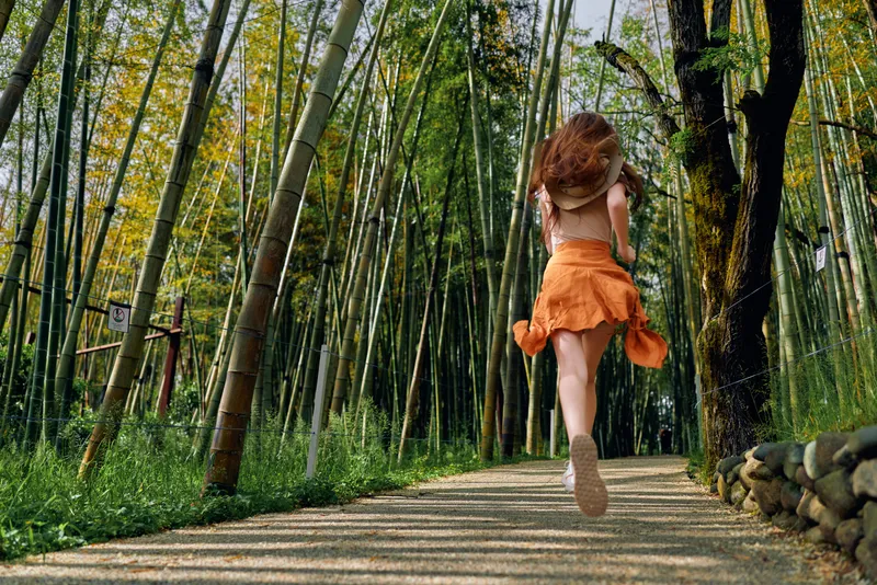 Woman running along a bamboo forest path in an orange dress, motion blur and back view among tall trees and nature, carefree jump on sunlit trail for outdoor adventure