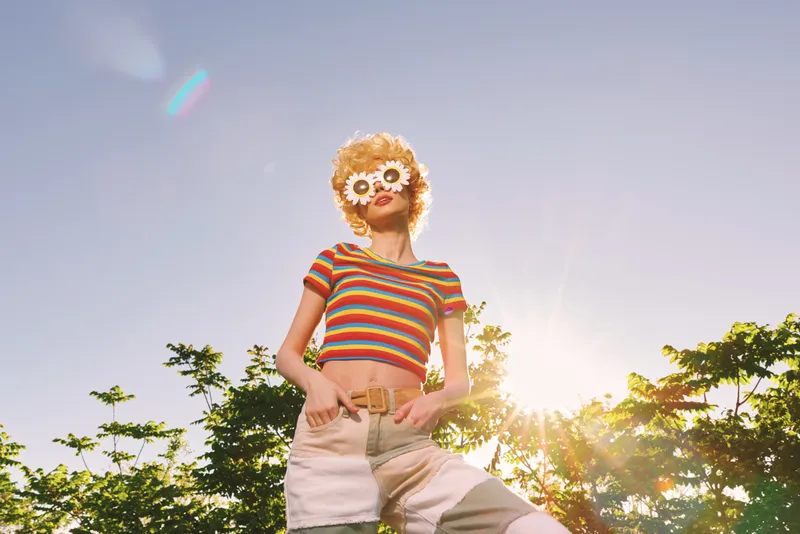 A woman stands outdoors in bright sunlight wearing a striped crop top and light shorts, a flower mask over her eyes. Bold retro color tones evoke carefree summer vibes.