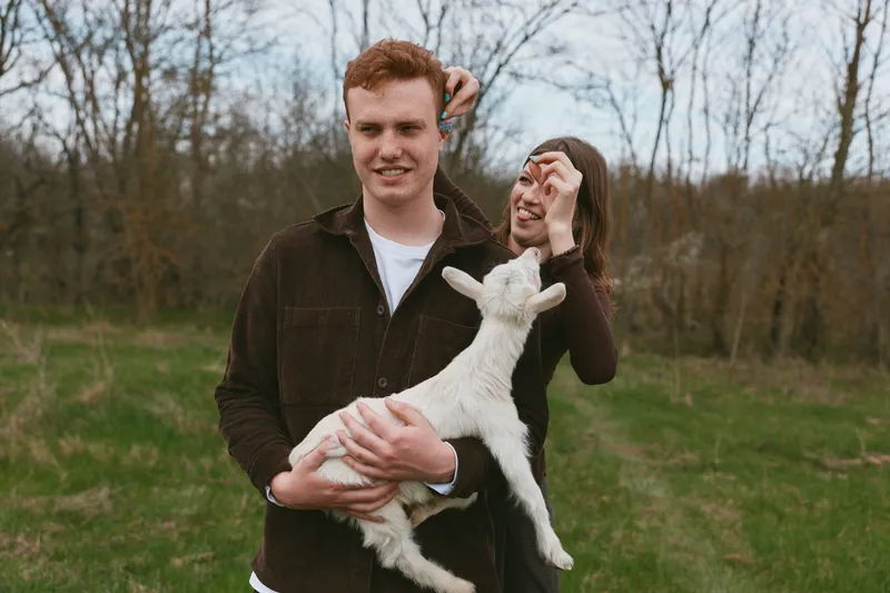 Smiling couple outdoors holds a playful goat kid, capturing a wholesome lifestyle moment in a grassy park under soft daylight, warm natural lighting emphasizing connection, care, joy, and simple