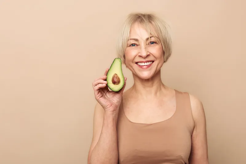 Smiling middle aged woman holds fresh avocado half near face on beige background. Concept for healthy eating, natural nutrition, wellness, vegan lifestyle, and positive mindset.