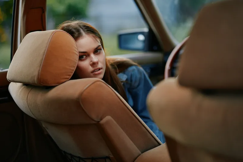 A young woman with long brown hair rests her head on a car seat, conveying a sense of contemplation and tranquility in a natural outdoor setting.