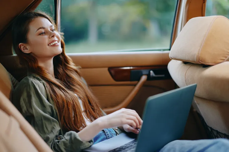 Young woman with long hair working on a laptop in a car, smiling, showcasing a relaxed and creative lifestyle in a natural setting.