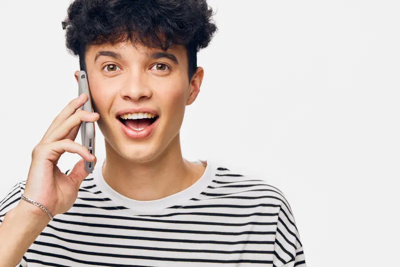 young man talking phone smiling, casual striped shirt, curly hair, joyful expression, communication, white background, studio light