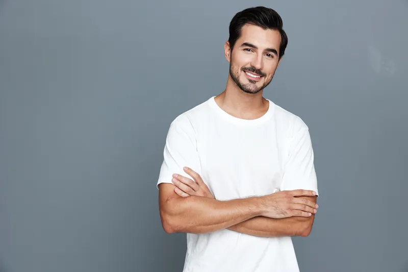 Smiling young man with dark hair and beard standing confidently with arms crossed against a plain grey background. Modern style and positive energy concept.