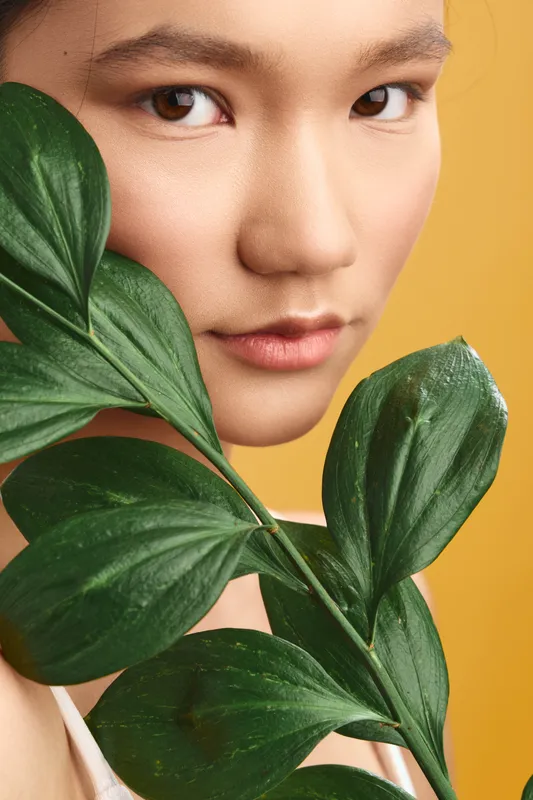 asian woman appears in a close up portrait, with large green leaves framing her face against a warm yellow backdrop, creating a fresh, serene and elegant mood.