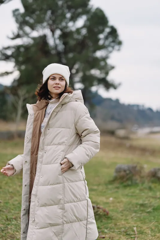 Woman outdoors in a long beige winter coat stands in an open field with distant trees, capturing a calm landscape and cozy outerwear perfect for chilly day photography