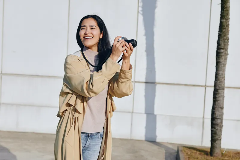 Camera in hand, a smiling woman captures candid outdoor moments, showcasing fashion and daylight as she enjoys a relaxed photography session beside a modern wall