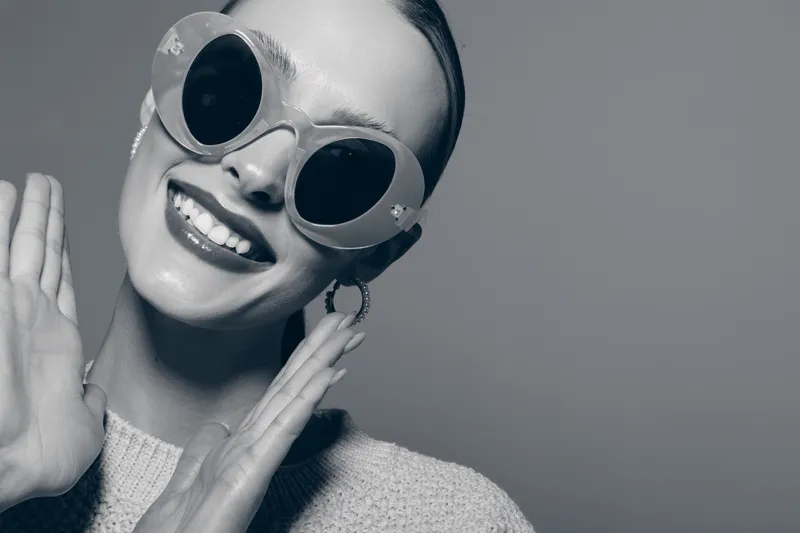 Smiling young white woman with pale skin wearing large round sunglasses and light sweater gestures happily isolated on grey background. Concept of joy, fashion and casual lifestyle.