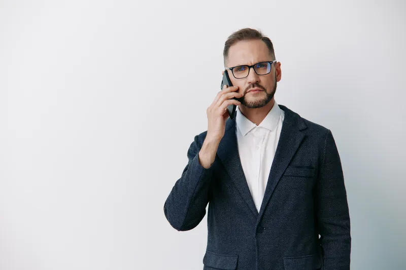 A business professional man speaks on a phone, standing against an isolated colored background, conveying confident focus and thoughtful emotion in a sleek corporate portrait.