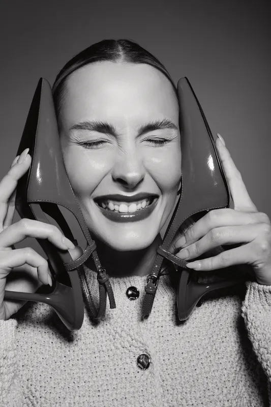 Young white woman with fair skin holds red high heels near her face with closed eyes and bright smile, wearing knitted sweater. Concept of happiness, fashion, and female style.