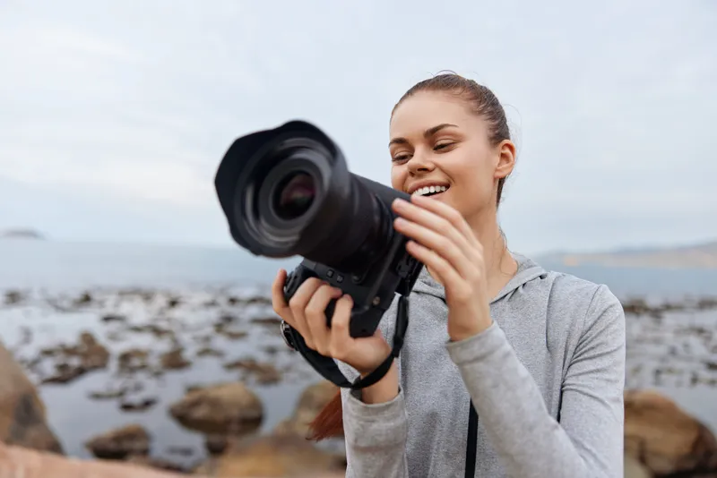 Young woman smiling while holding a camera by the seaside, wearing a gray hoodie, showcasing a joyful expression, surrounded by rocks and water