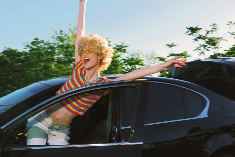 Energetic woman with a curly blonde wig leaning out of a sleek car window, striped shirt, vibrant retro color palette, carefree street style, sunny day, dynamic motion.