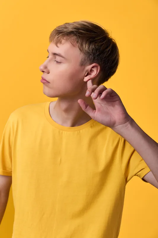 Youthful man in a yellow tshirt touches ear with a calm expression, listening in a minimalist studio with vibrant backdrop, casual fashion, sideview