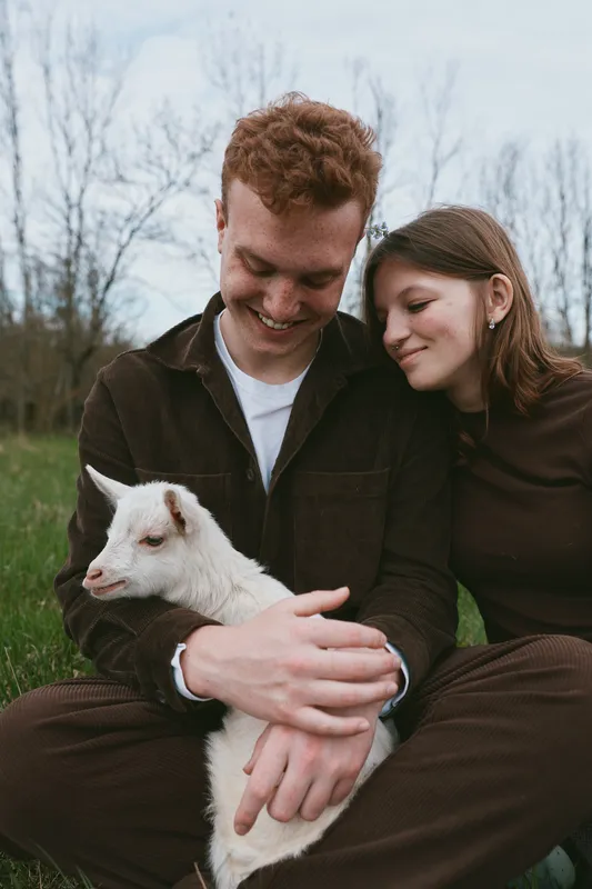 Candid couple outdoors gently cradle a young goat, sharing a warm, relaxed moment among green fields, soft light, and fresh breeze, capturing affectionate companionship and wholesome rural lifestyle