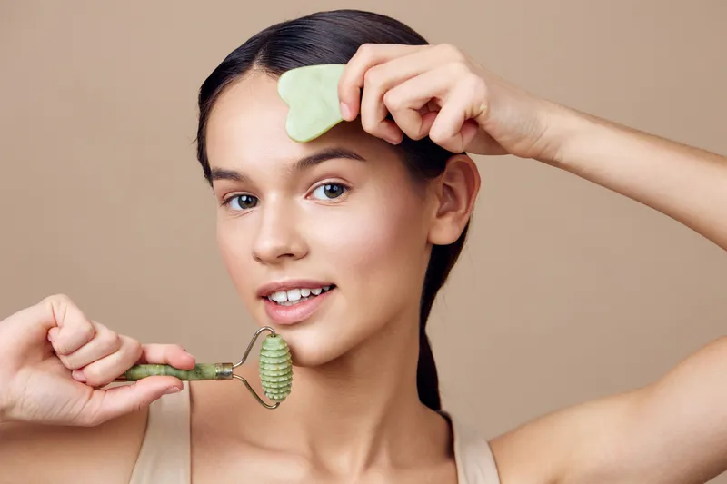 Young woman enjoying a skincare routine with a jade roller and gua sha tool, showcasing healthy skin and relaxation in a serene beige background