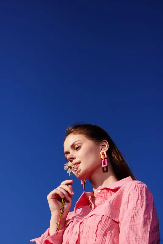 Young woman with pink shirt enjoying a flower under a clear blue sky, reflecting joy and positivity, perfect for springtime themes and healthy living concepts