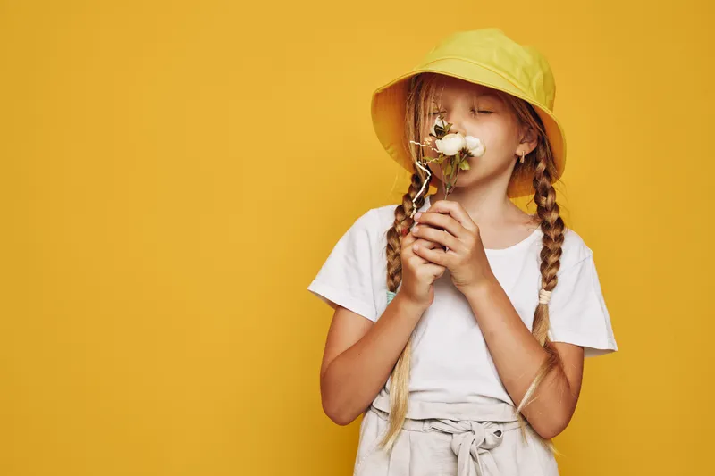 Young girl in a yellow hat with braided hair sniffs a white flower against a yellow background. She wears a white shirt and beige pants. Concept of nature and childhood.
