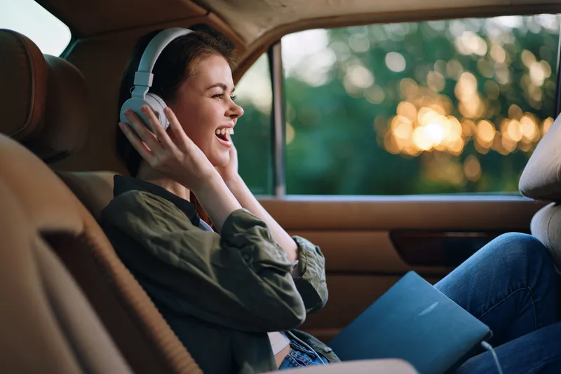 Joyful young woman enjoying music in a car, wearing headphones and smiling with a sunset background, capturing happiness and leisure.