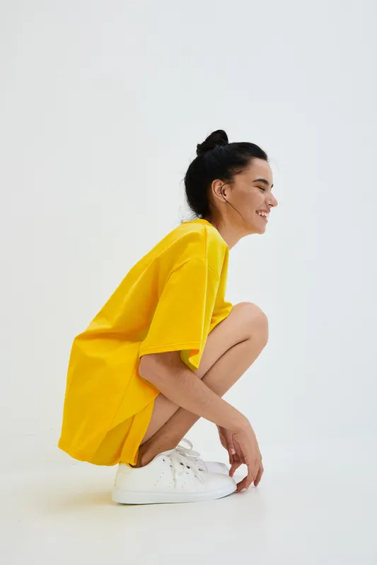 smiling woman wearing bright yellow oversized t-shirt and white sneakers crouching on a white seamless background in studio, casual style and joyful mood