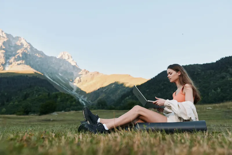 A cheerful young woman sits on a mat in a scenic meadow, typing on a laptop. She smiles and appears focused, creating a calm and productive outdoor work atmosphere.