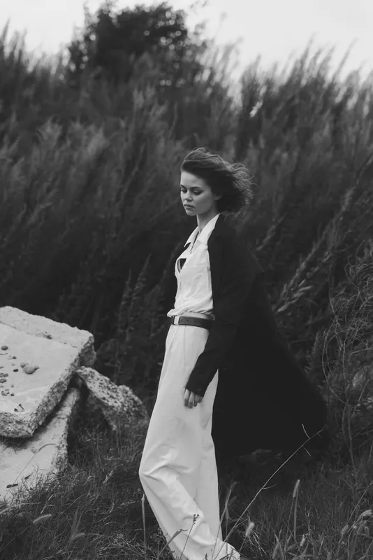 Woman in flowing dress and cloak stands among rocks and grass, wind sweeping through hair, a monochrome scene conveying solitary grace, nature and movement in a moody outdoor landscape
