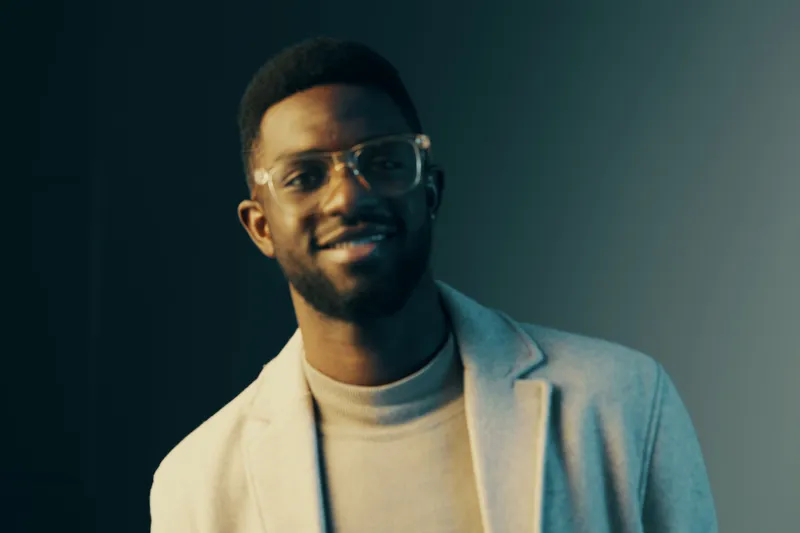 Young Black man wearing glasses and a stylish suit, smiling confidently against a gradient background, representing modern fashion and professionalism
