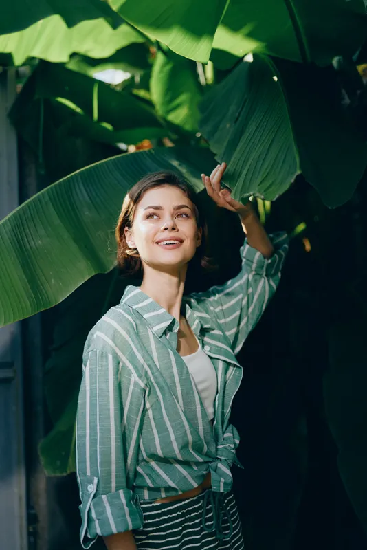Portrait of a smiling woman among lush tropical leaves, wearing a striped shirt in soft sunlight.