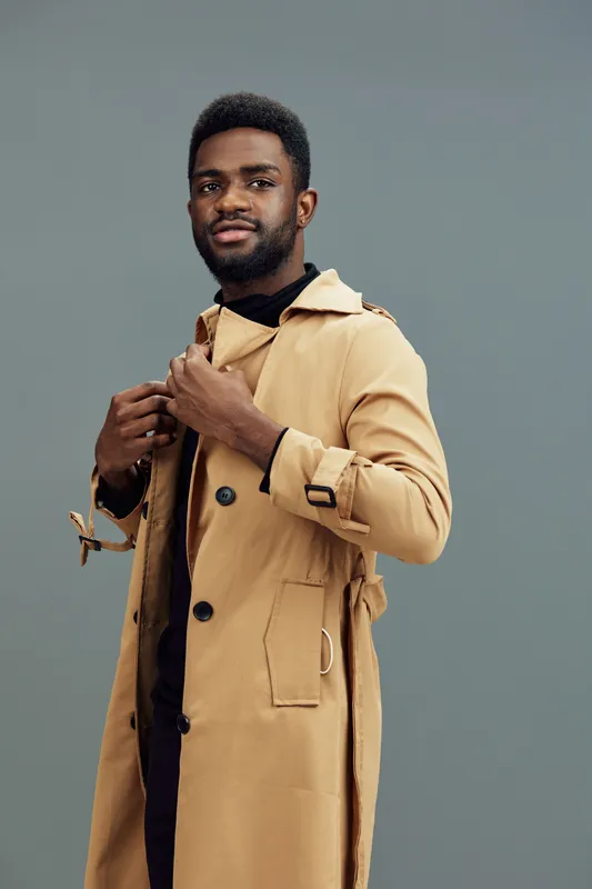 Stylish young Black man wearing a beige trench coat against a muted backdrop, exuding confidence and sophistication during the fall season