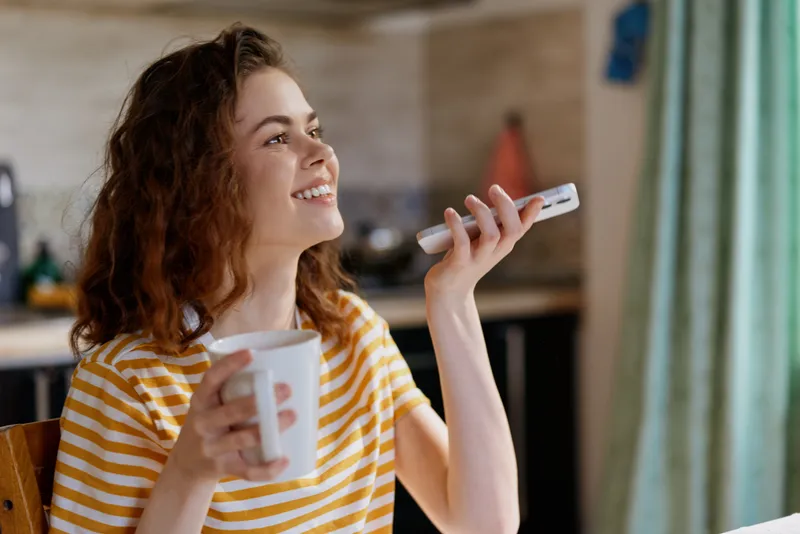 A smiling woman in a striped shirt sits at home, holding a mug in one hand and a phone in the other. A cozy, sunlit morning scene capturing relaxed domestic life.