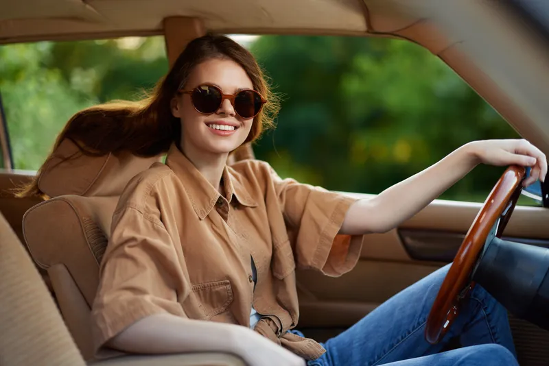 Young woman smiling in a vintage car, wearing sunglasses and a casual brown shirt, capturing a joyful summer vibe with green nature in the background.