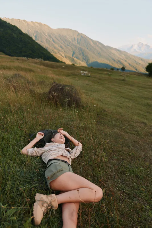 A woman lying on grass in a scenic valley with distant mountains, enjoying a quiet moment outdoors. A lifestyle shot focused on relaxation, nature, and well being.