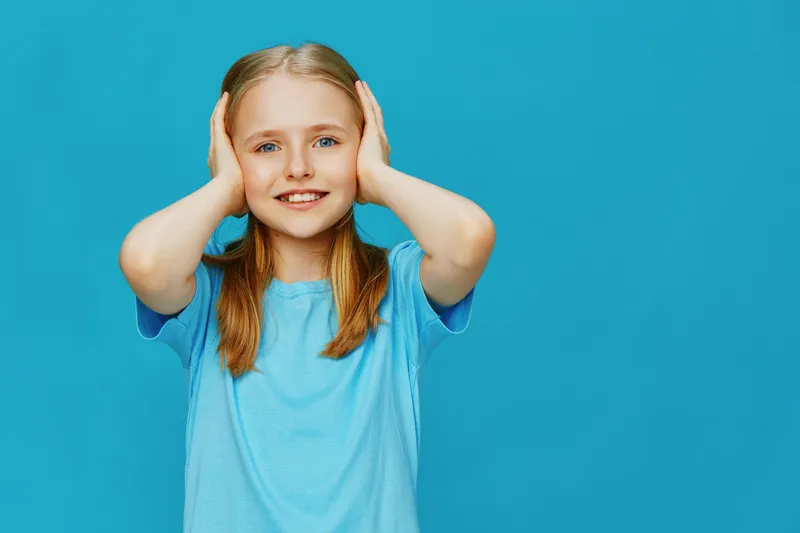 Cheerful young girl covering her ears against a bright blue background, wearing a light blue t shirt She expresses joy and playfulness, perfect for children s themes