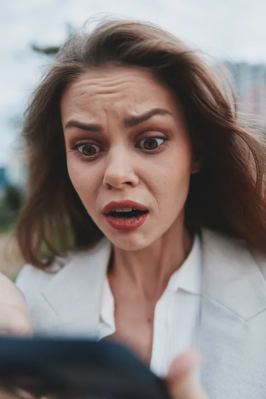 Woman shocked by financial crisis looking at phone, upset young Caucasian female in business attire outdoors with blurred city background, anxious emotion, stock market drop theme
