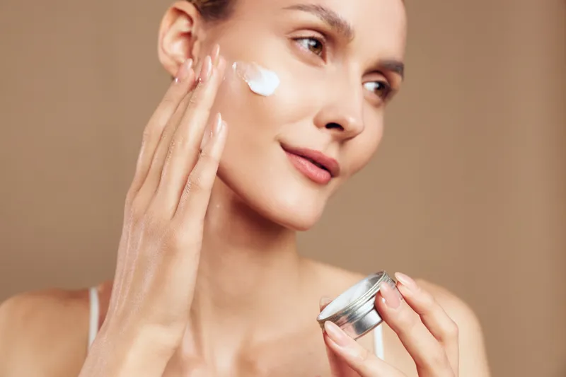 Young woman applying moisturizer cream on face, smooth skin care routine, natural makeup, gentle touch, soft lighting, neutral background, healthy beauty concept