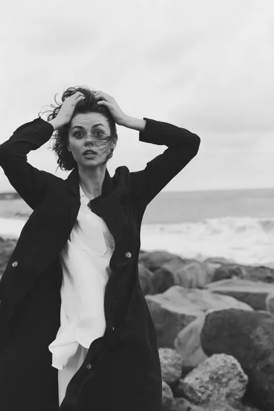 Moody black and white beach scene with wind swept coat, hair and mist across face, dramatic fashion photography on a rocky shore against windy horizon