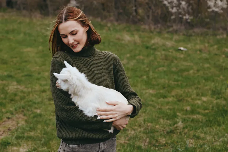 Young woman in a green sweater gently holds a white baby goat outdoors on a grassy field, smiling softly while enjoying a peaceful moment with the animal in a natural setting.