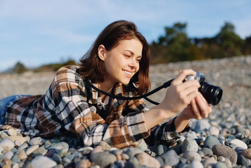 a woman photographer lies on a rocky beach using a camera to capture the scene casual plaid shirt pebbles around bright day focused expression creative moment in outdoor nature photography