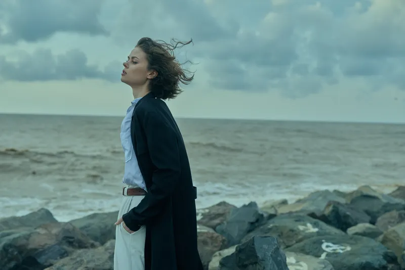Woman standing on a rocky coastline wearing a long dark coat and light shirt, hair blown by wind, facing the sea with a calm, contemplative mood, cloudy sky and open water