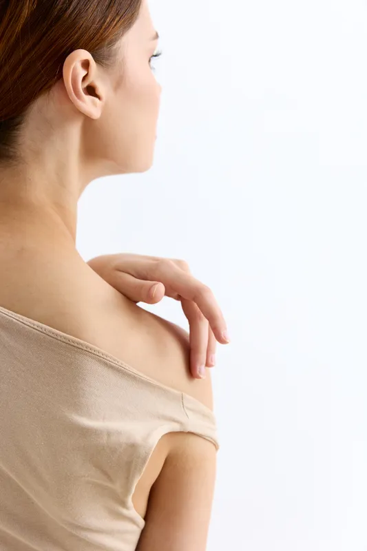 woman touching shoulder with gentle hand in soft beige top showing delicate skin and calm posture on light background, minimalistic beauty concept