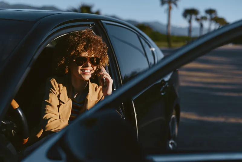 A stylish woman sits in a dark car, wearing sunglasses and a tan blazer, smiling while on a call. Bright sunlight and a sense of travel capture a casual road moment.