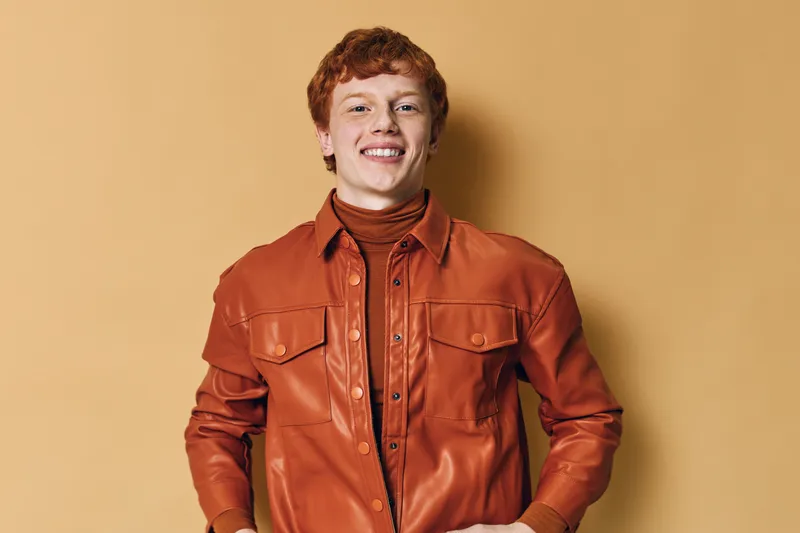 Young man with red hair wearing a brown leather jacket smiling confidently on beige background. Portrait of cheerful person with casual style and relaxed posture indoors.