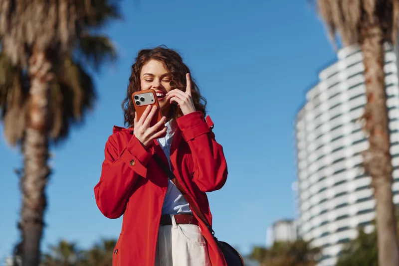 Candid woman outdoors in a red coat, smiling while talking on a phone, sunlit urban street scene with palm trees and modern buildings in bright sunshine.