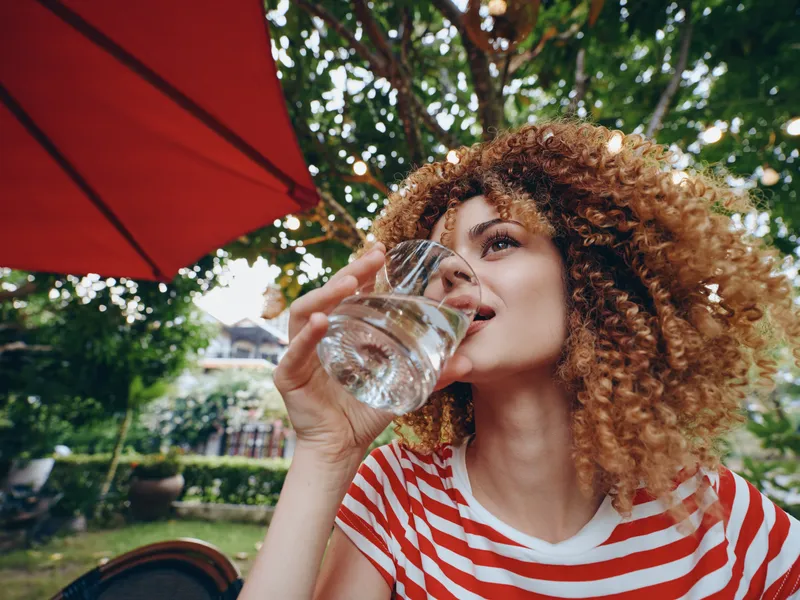 A woman with curly hair in a red striped shirt drinks water from a glass outdoors, surrounded by lush greenery and a garden patio, capturing a refreshing moment.