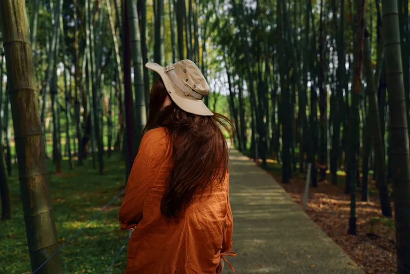 Woman back walking on path through bamboo forest wearing a hat and orange coat, longhair flowing. Scenic walkway with trees and sunlight, peaceful outdoor travel and contemplative mood.