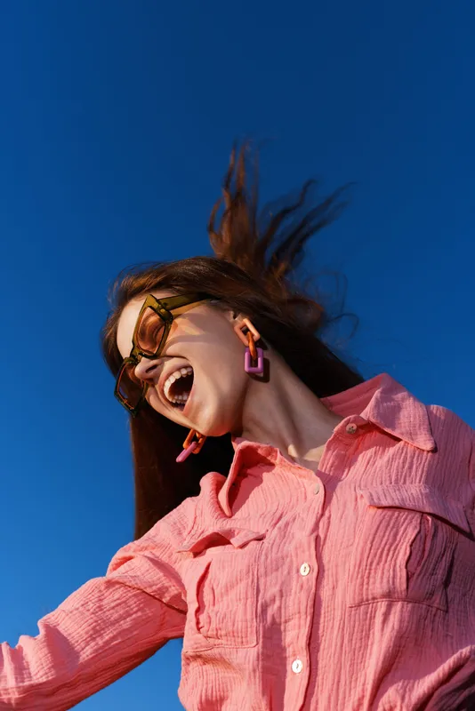 Excited woman expressing joy outdoors in a vibrant pink shirt with large earrings against a clear blue sky, capturing a lively and carefree summer vibe