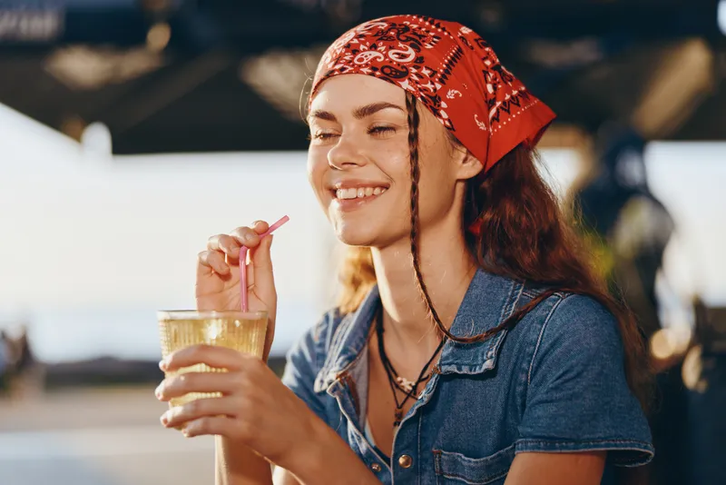 smiling woman with red bandana enjoying a cold drink outdoors, casual denim shirt, warm sunlight, relaxed summer atmosphere, cheerful expression