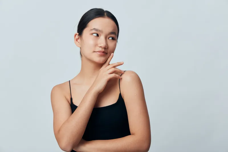 Young woman with smooth skin and dark hair in black tank top looking sideways with thoughtful expression on plain light background. Portrait, beauty and fashion concept with minimal style.