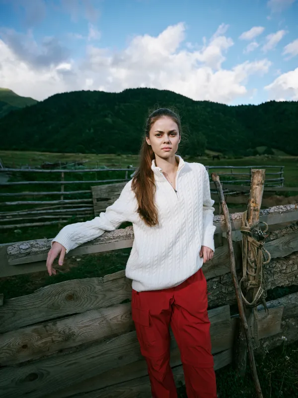 Woman leaning on a weathered wooden fence in a mountain meadow, wearing a white sweater and red pants, calm expression, expansive sky and green hills in a rural outdoor setting