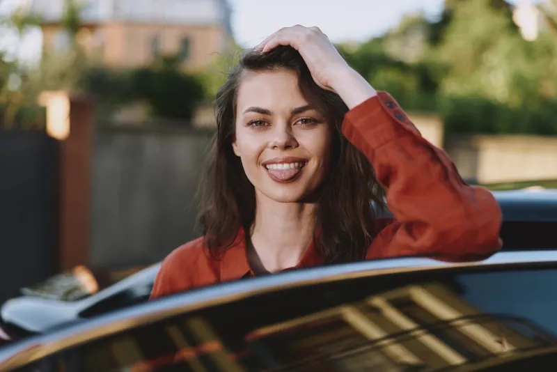 A cheerful woman leans out of a car window, wind in her hair, enjoying a sunny day. A lifestyle portrait that communicates confidence, freedom, and relaxed urban moments